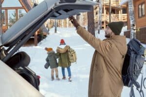 Young man with big rucksack shutting down cover of car trunk while standing against his wife and daughter moving towards country house - arriving in the snow - road trip - getaway