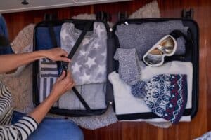 Top view of unrecognizable woman sitting on wooden floor and fastening a belt on opened suitcase above warm clothes near ski goggles