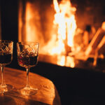 Two glasses of red wine on wooden table with cozy fireplace flame on the background. Winter season