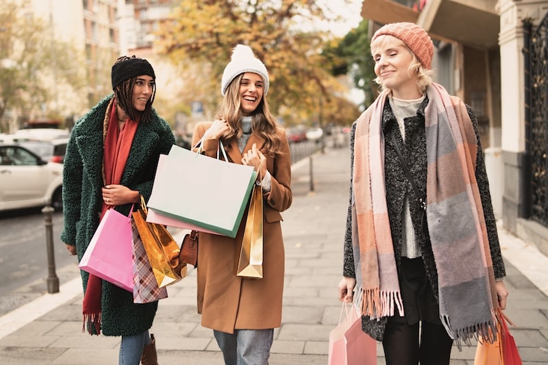 Three young women stroll through city streets, smiling and chatting happily with numerous colorful shopping bags in hand, immersed in the vibrant autumn atmosphere.