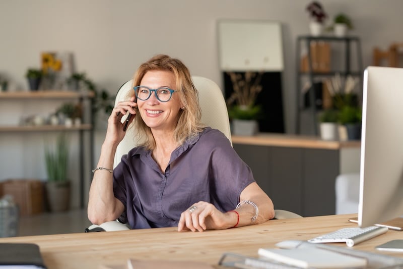 Happy mature blond businesswoman in casualwear sitting by desk in front of computer in living-room while talking on smartphone