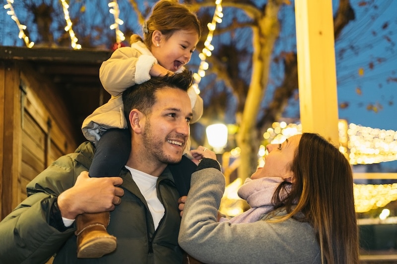 Young father holding his young smiling daughter sitting on his shoulders next to her mother as they visit a city decorated for Christmas. Concept of happy two-parent heterosexual family.