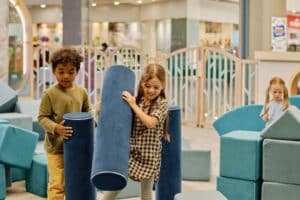 Smiling little girl in checkered shirt carrying big soft toy roller of blue color while building tower or house with cute boy
