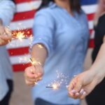 three unidentifiable women holding sparklers in font of an American Flag 4th of July