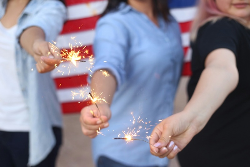 three unidentifiable women holding sparklers in font of an American Flag 4th of July