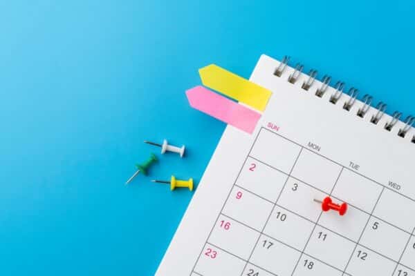 A top-down view of a spiral desk calendar with colorful arrow sticky notes and push pins, with one red pin marking a specific date. Planning, scheduling, and time management concepts.